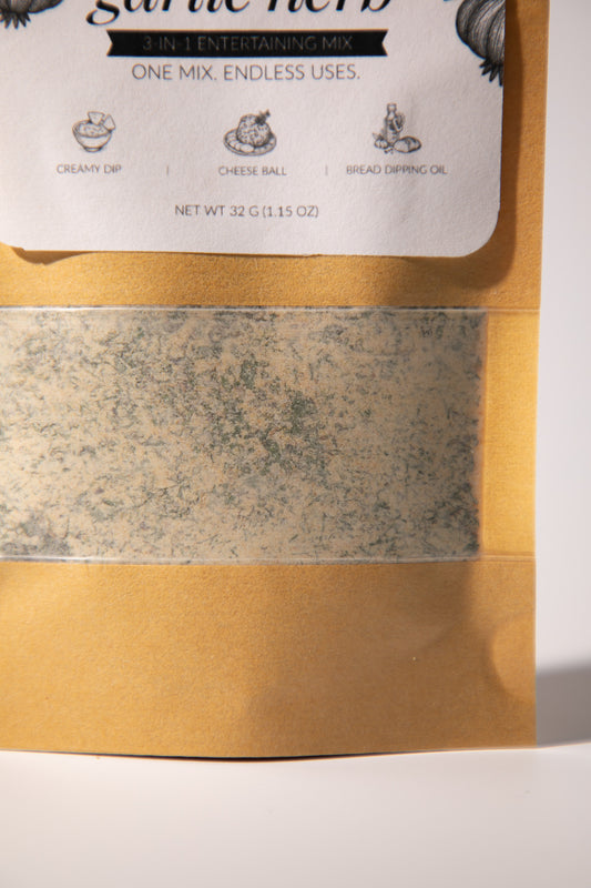 A close-up of a garlic herb seasoning mix in a kraft paper pouch with a clear window showing the textured herb blend inside. The white label at the top reads 'garlic herb' and lists uses like creamy dip, cheese ball, and bread dipping oil.